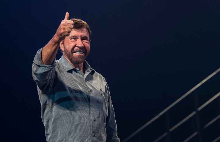 Chuck Norris sorride mentre fa il gesto dell'ok col bracccio alzato con dietro a sé uno sfondo scuro.