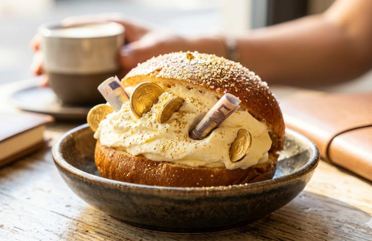 Un delizioso maritozzo romano con panna montata, spolverato con polvere d'oro e zucchero e con banconote e monete incastonate al suo interno, servito su un tavolo di legno illuminato dal sole in un caffè italiano. Rappresentazione dei consumi della Mood Economy nel 2026.