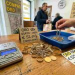 Un'inquadratura ravvicinata del bancone di un negozio Compro Oro in Italia, con una bilancia digitale che mostra il peso di diversi gioielli d'oro. Sulla superficie di legno, un cartellino con la scritta "In Calo!" evidenzia il trend del mercato. Sullo sfondo, un cliente e un orologio da parete.