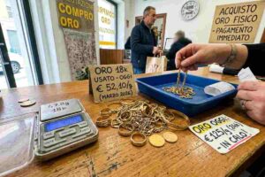 Un'inquadratura ravvicinata del bancone di un negozio Compro Oro in Italia, con una bilancia digitale che mostra il peso di diversi gioielli d'oro. Sulla superficie di legno, un cartellino con la scritta "In Calo!" evidenzia il trend del mercato. Sullo sfondo, un cliente e un orologio da parete.