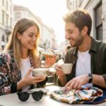 Giovane coppia felice che sorride e si gode un maritozzo con panna e caffè su un tavolo di legno illuminato dal sole in un bar italiano. Esempio dei consumi della Mood Economy nel 2026.